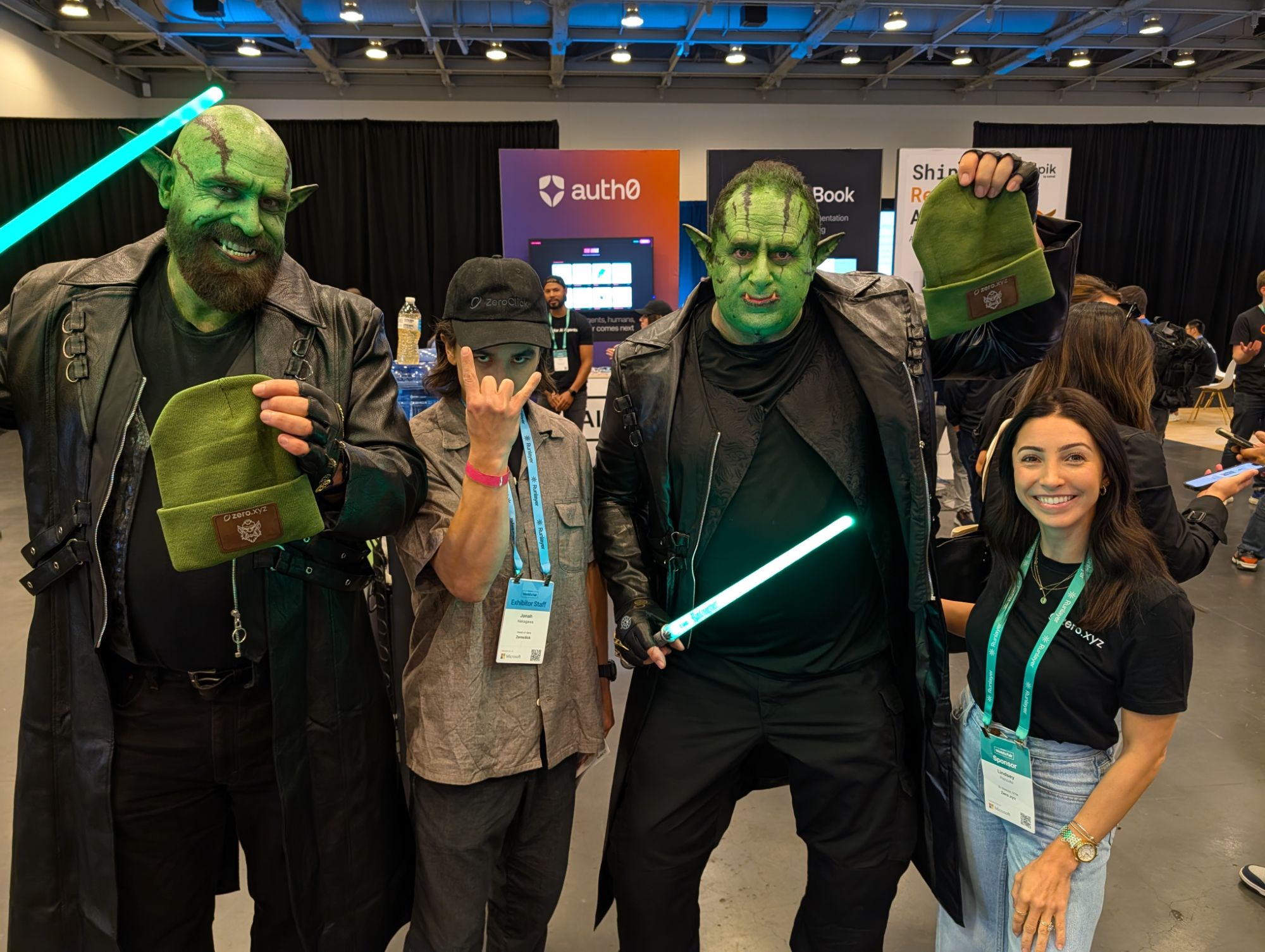 Conference guests in orc costumes holding up their green beanies with engraved patches, posing with booth staff